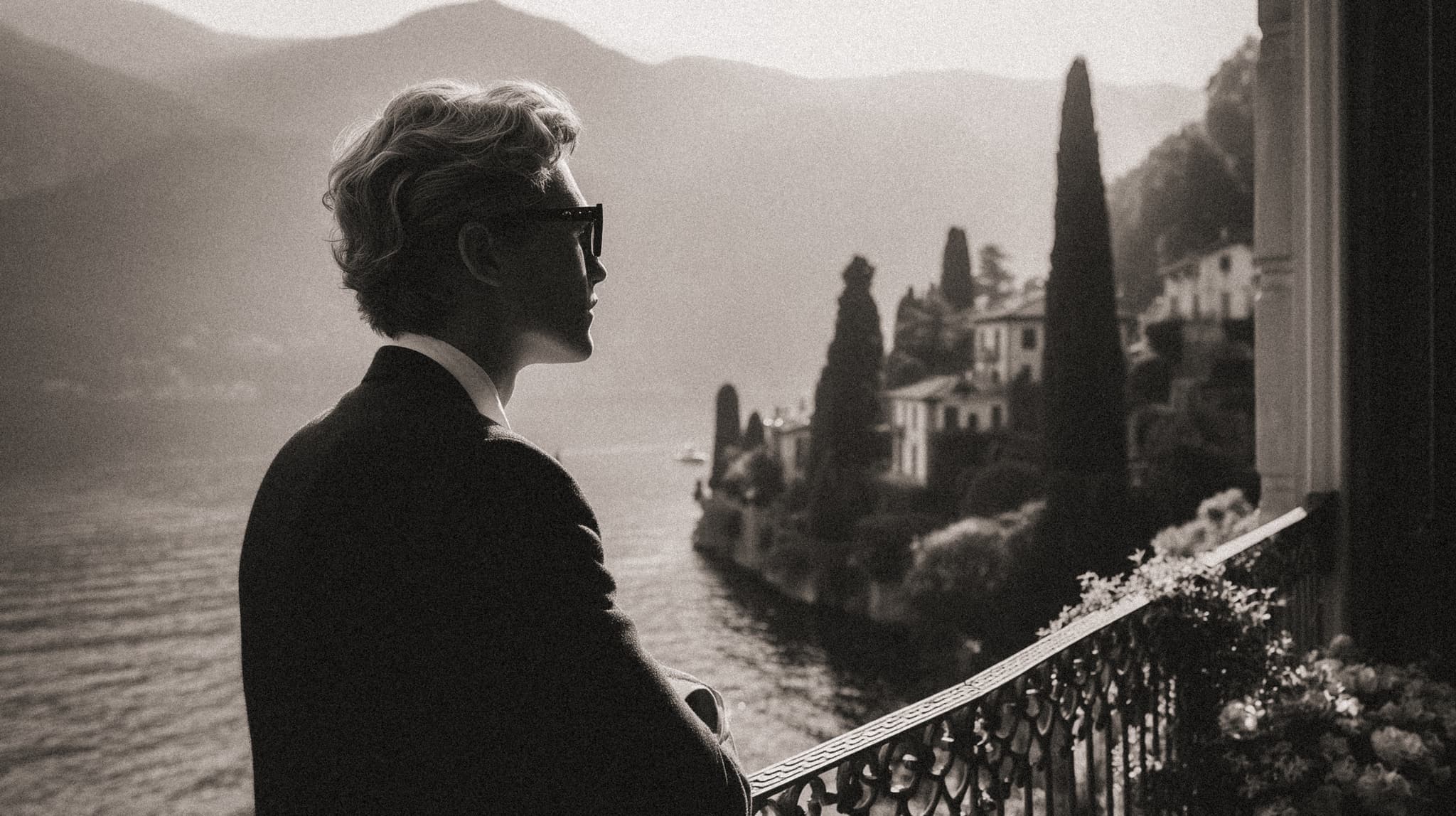 Back view of a man in tailored suit and sunglasses on a villa balcony with Lake Como and mountains in the distance.